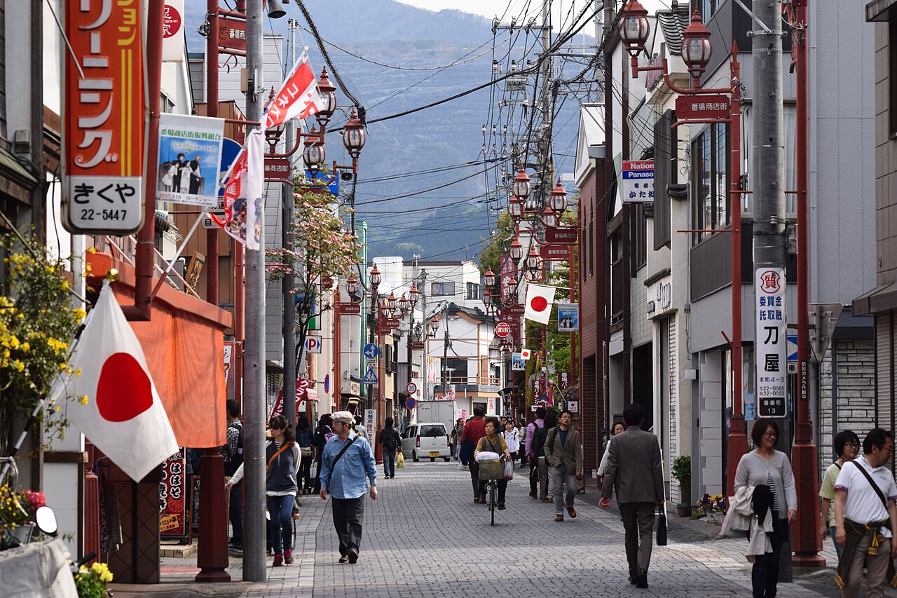 The Bamba shopping street in central Chichibu, Saitama on an ordinary afternoon: a paved lane lined with low shops, red-painted street lamps and hanging flags and banners, pedestrians and a cyclist moving at walking pace, and a wooded mountain ridge closing off the far end of the street.