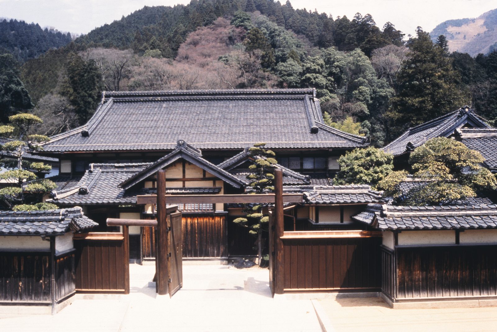 Open wooden gate and the large tiled roof of the Ishitani residence in Chizu, with wooded hills behind