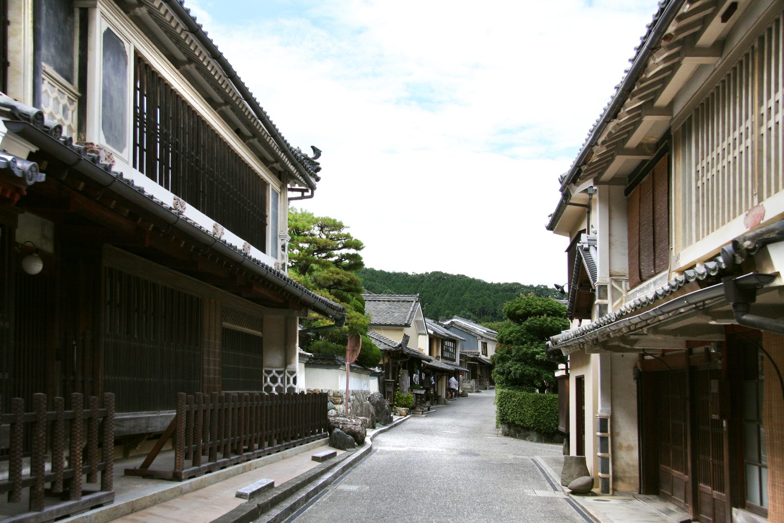 A narrow paved lane running between old merchant houses with latticed windows and tiled eaves in the Yokaichi-Gokoku historic district, with a forested hill beyond