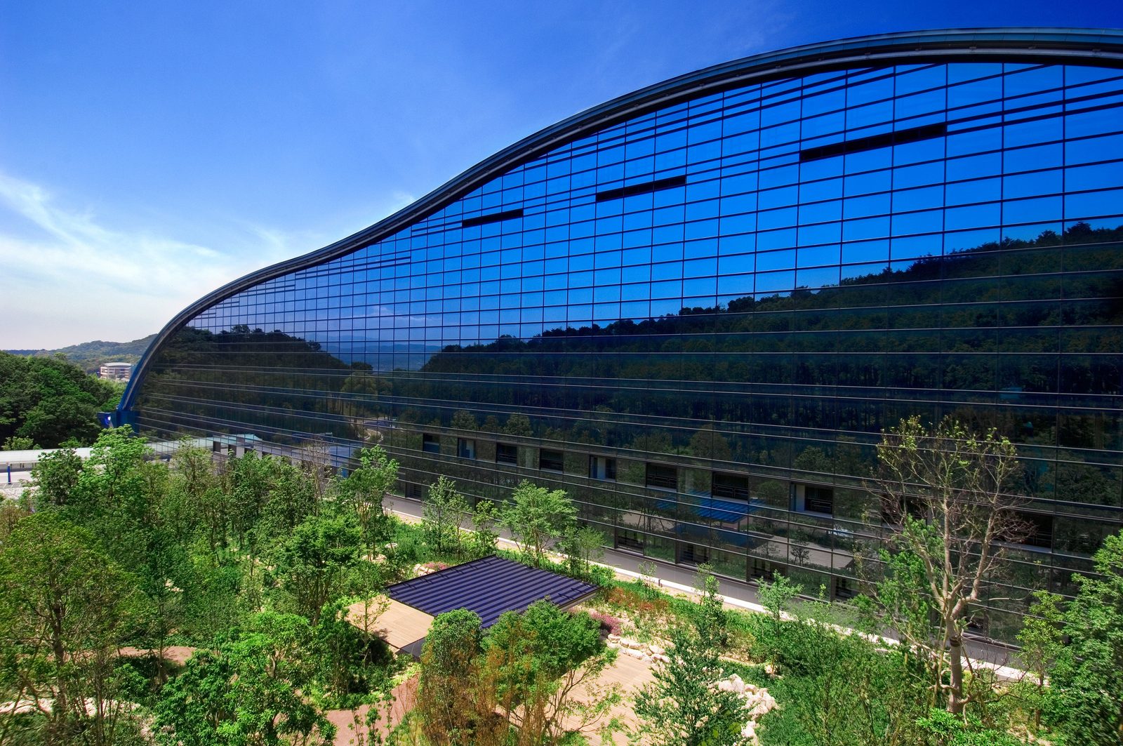 The curved glass exterior of the Kyushu National Museum reflecting the green mountains behind it.