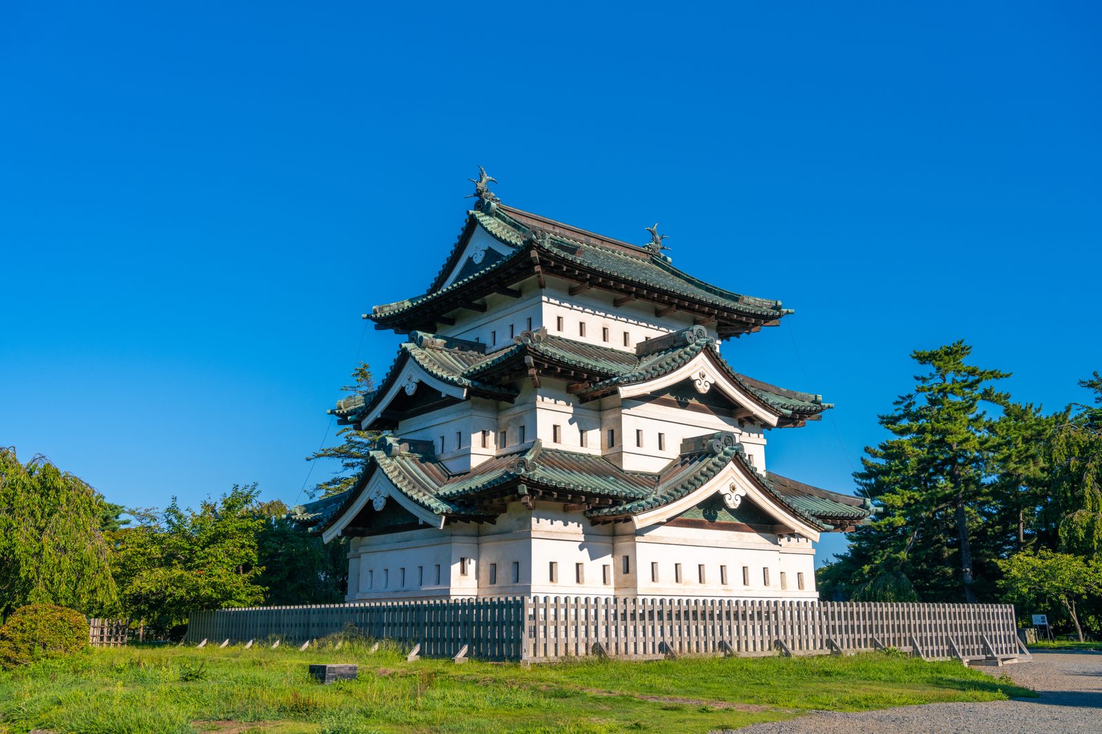 The three-storey keep of Hirosaki Castle, white plaster above a stone base with green tiled roofs, standing behind a low wooden fence under a clear blue sky.