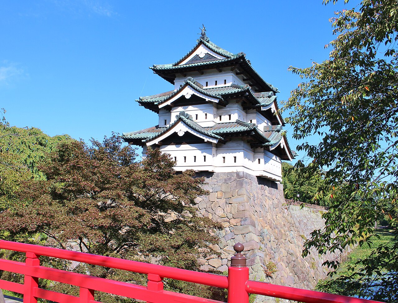 The white three-storey keep of Hirosaki Castle on its stone base, seen from a red-railed bridge with green and reddening foliage around it under a clear sky.