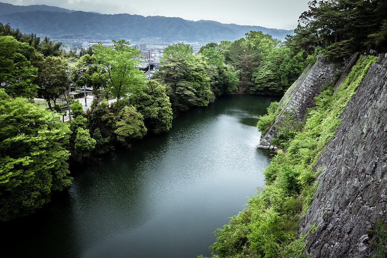 A moat of deep green water below the towering stone walls of Iga Ueno Castle, with trees and distant mountains beyond