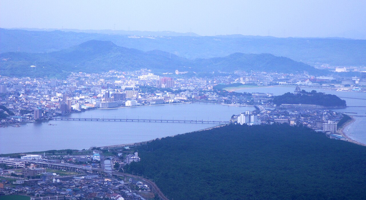 An elevated view over central Karatsu, with the Matsuura River, a bridge, and the old town's rooftops stretching toward the harbor and Niji-no-Matsubara in the distance.