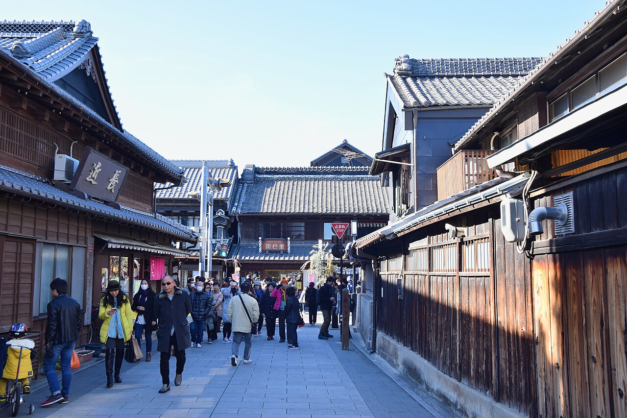 A street lined with traditional tiled-roof warehouse buildings in Kawagoe's old town, with visitors walking along the stone-paved lane.