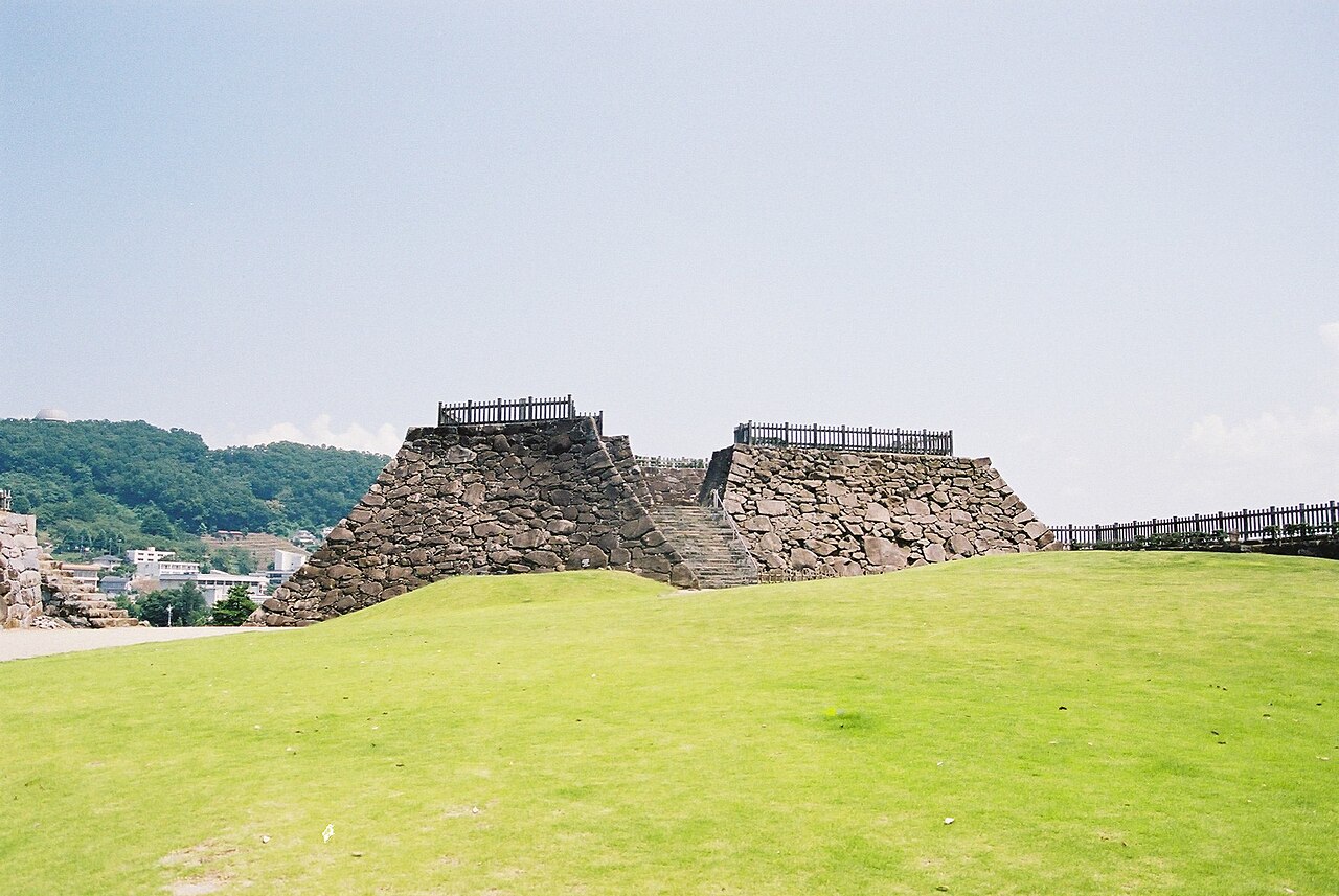 The empty lawn and stone keep base of Kofu Castle under a summer sky.
