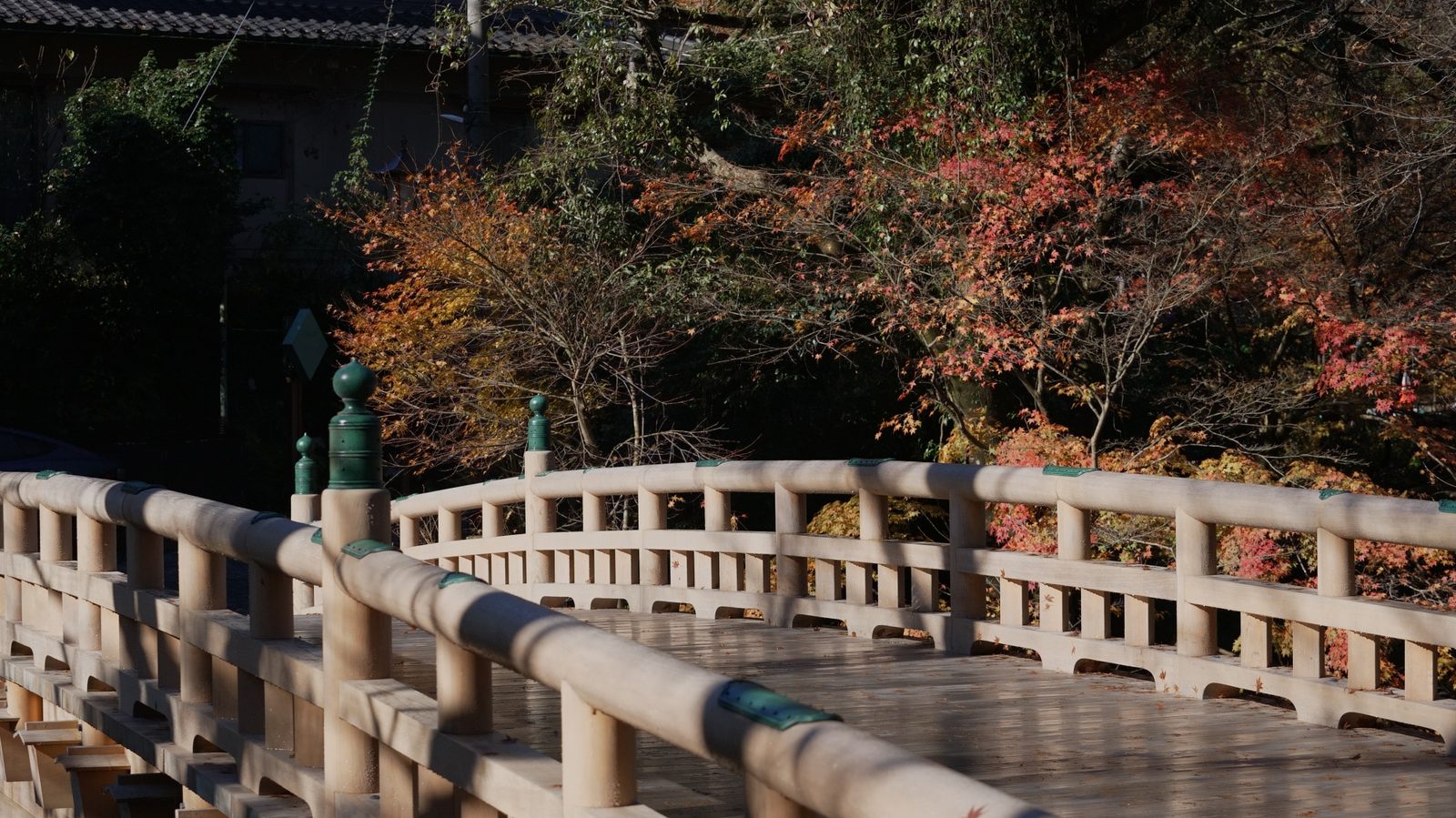 A pale wooden footbridge with rounded rails and green metal finials, backed by red and orange autumn maples, with the roof of a building visible above.