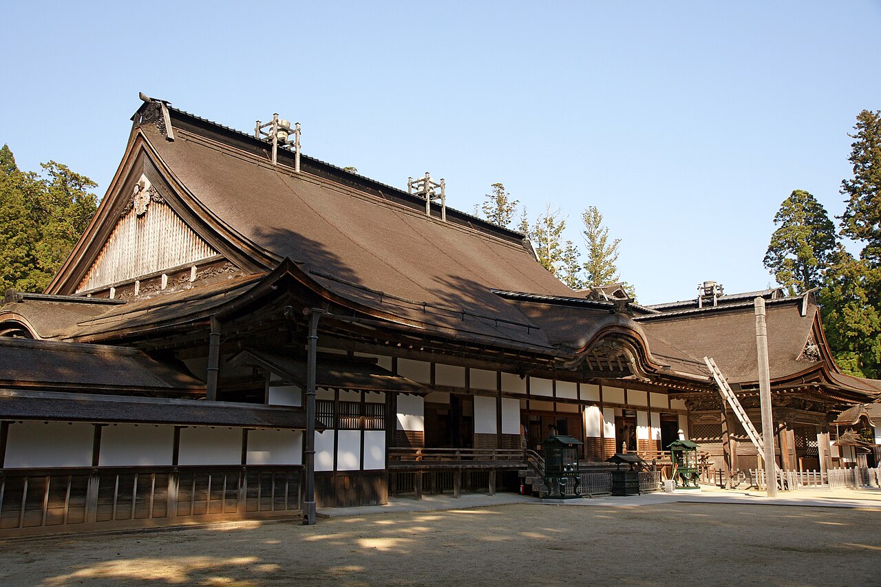 The wide cypress-bark roof of the main hall at Kongobuji sweeps across a bright gravel courtyard, its white plaster walls and dark timber verandas lit by low sun.