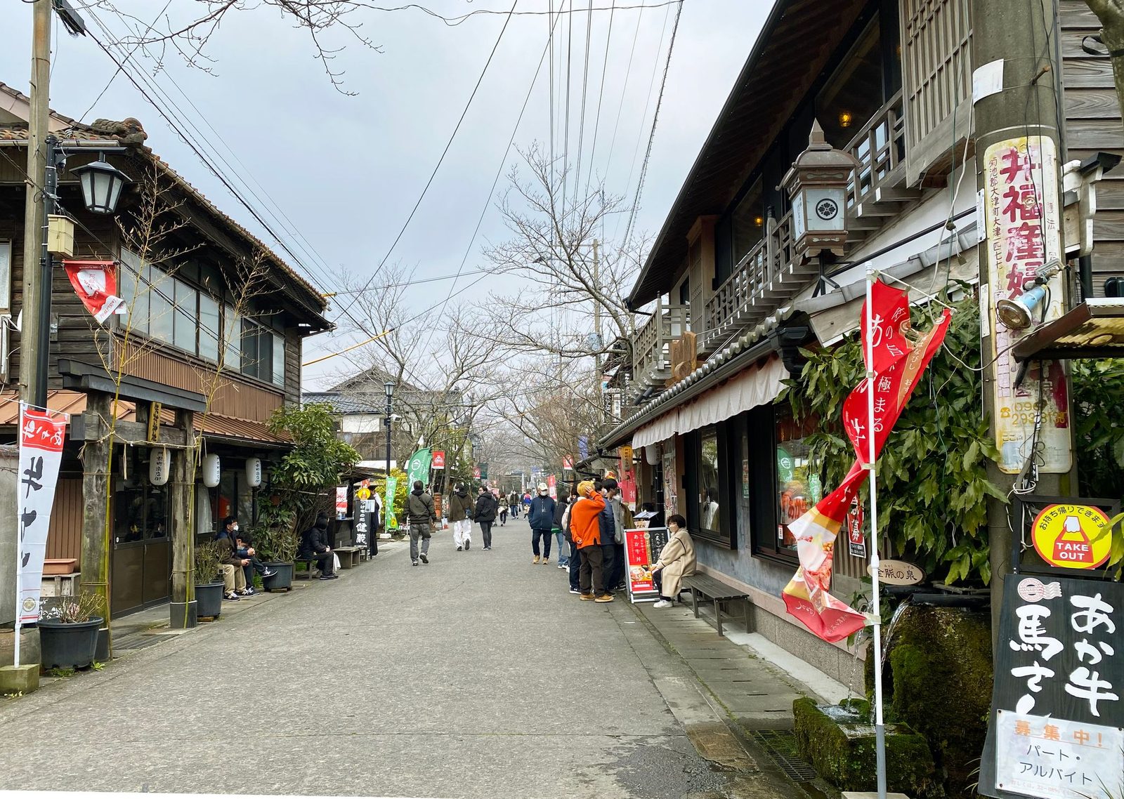 A street in the Aso Shrine monzenmachi with old wooden shops on both sides, red banners, bare cherry trees and people walking and queuing along the road.