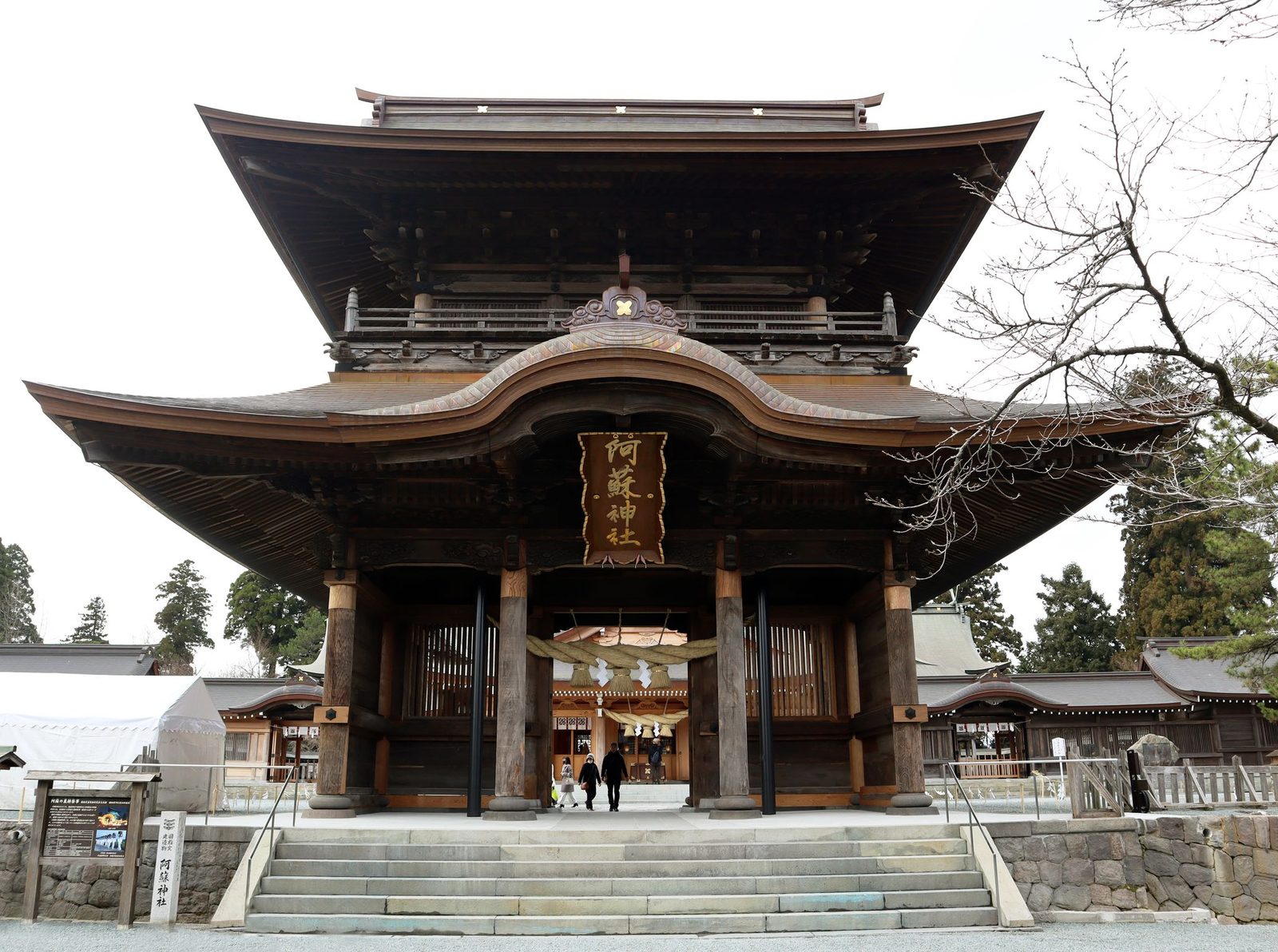 Front view of the two-story Roumon gate of Aso Shrine with its gold-lettered name plaque, a shimenawa rope under the eaves, and a few visitors walking through the gate.