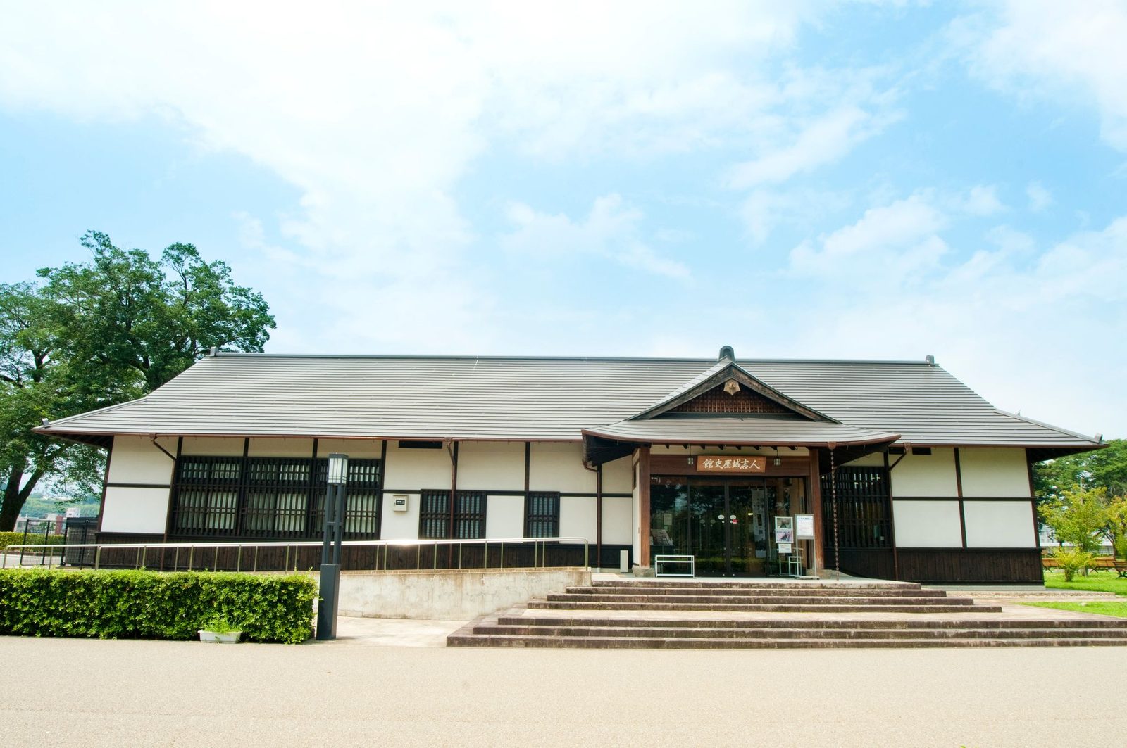 The white-walled, dark-timbered building of the Hitoyoshi Castle History Museum, with a gabled entrance porch and wide stone steps, under a blue sky with trees behind.