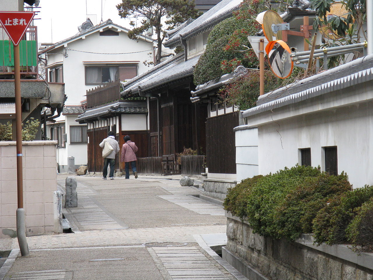 The former Sugiyama residence in Tondabayashi, a merchant house with dark wooden walls and tiled roofs beside a paved lane where two people are walking