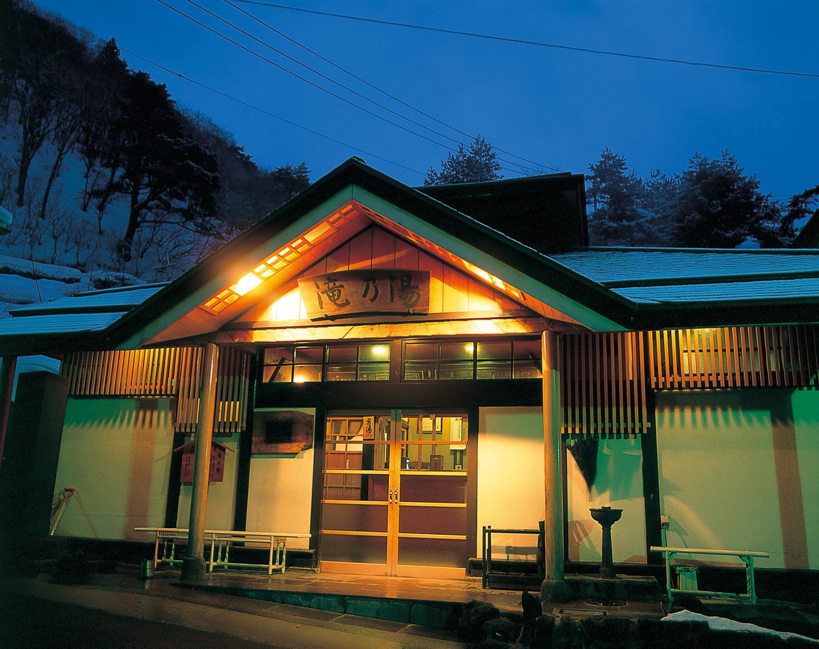 The wooden entrance of the Takinoyu public bathhouse in Naruko Onsen, its gabled roof lit from within above a carved name board, with snow on the roofs and a pine-covered slope behind.