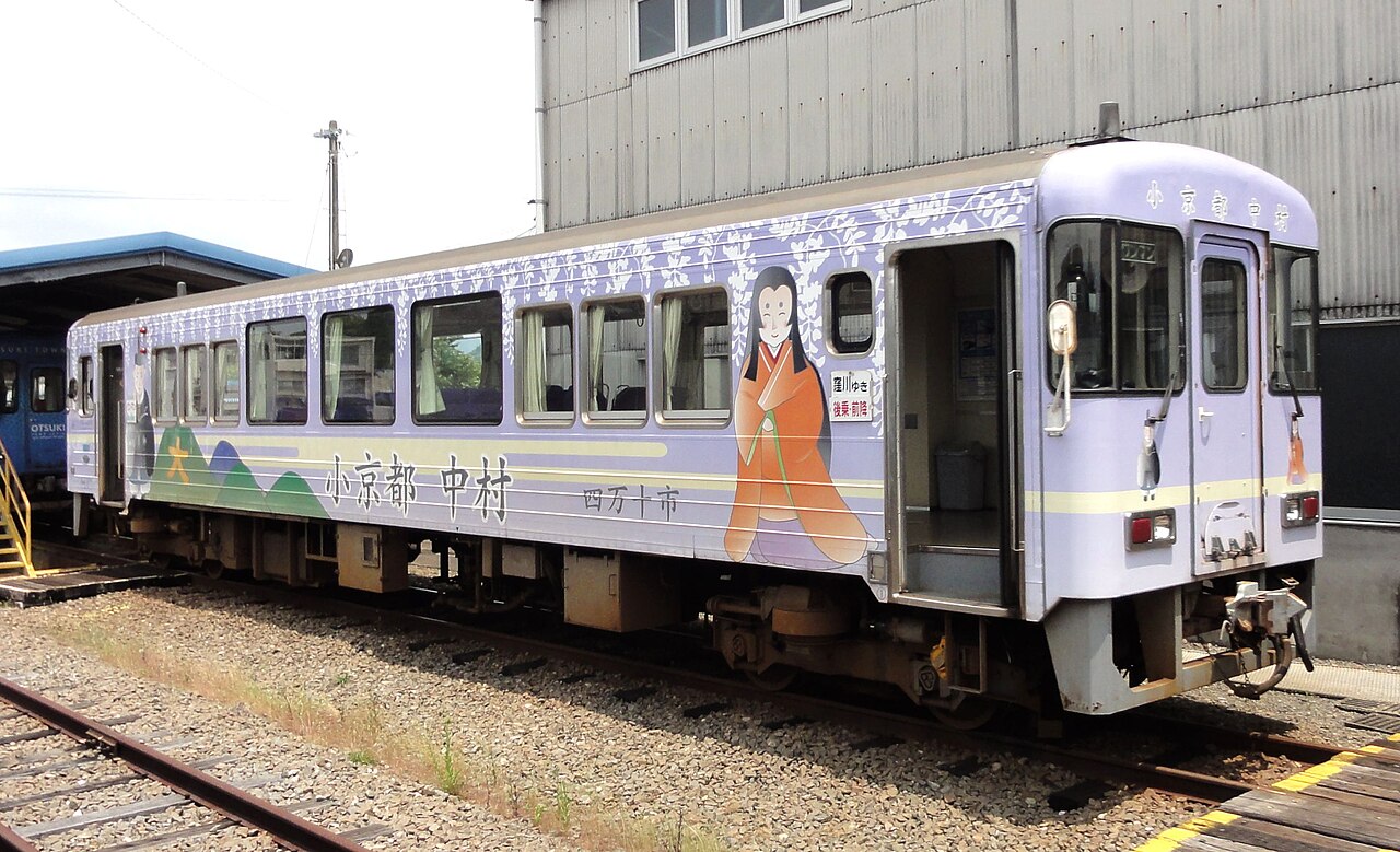 A single-carriage Tosa Kuroshio Railway train in pale purple livery, decorated with a Heian-period court figure and the name of Nakamura, standing on the track beside a depot building.