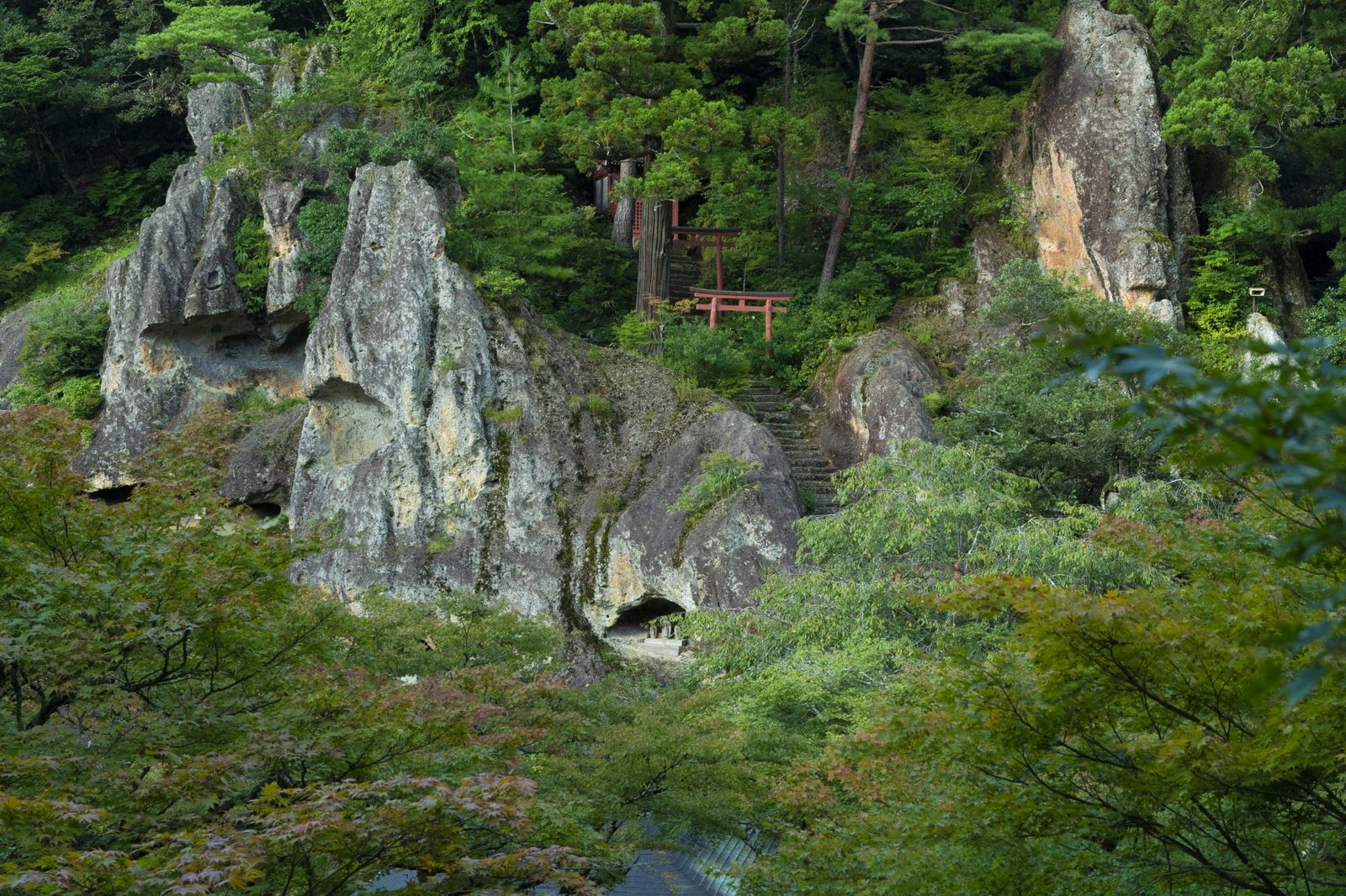 Weathered rock crags with cave-like hollows rise out of green forest at Natadera, with a small red torii gate and a stone stairway set into the slope.