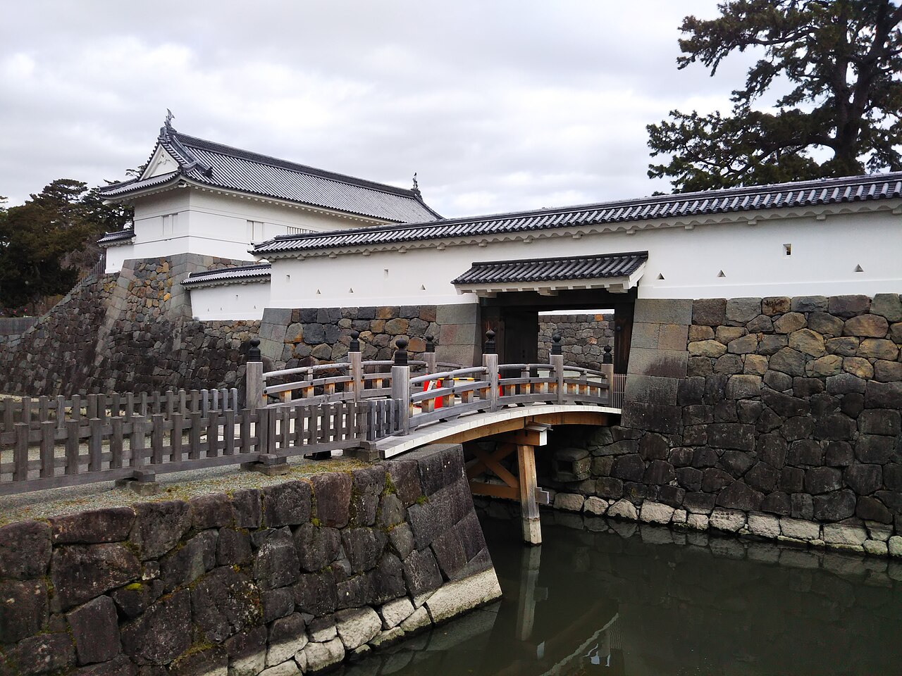 The arched wooden Sumiyoshi Bridge over the moat, leading to the white walls, stone base and turret of the Akagane Gate under an overcast sky.