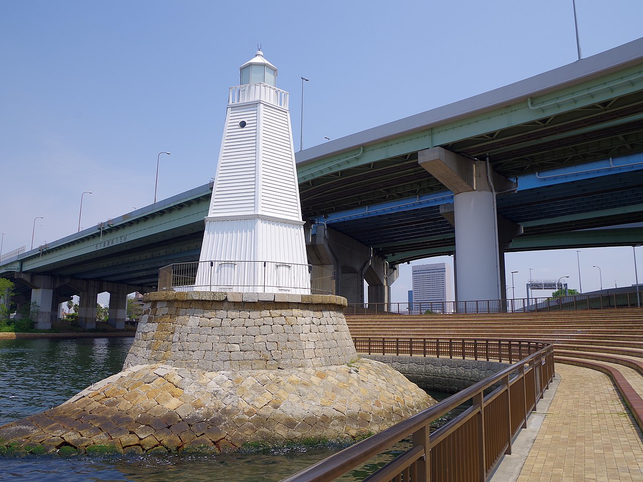 The white weatherboarded Old Sakai Lighthouse stands on a stone base at the water's edge, with an elevated expressway crossing behind it.