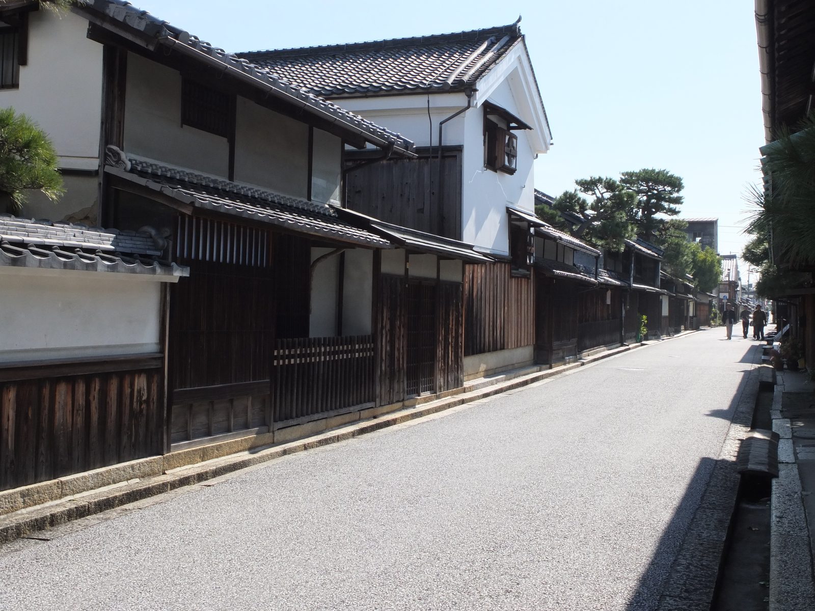 A quiet street of old merchant houses in Omihachiman, with dark timber lattice fronts, a white-walled building with a tiled gable, pine trees and a few pedestrians in the distance.