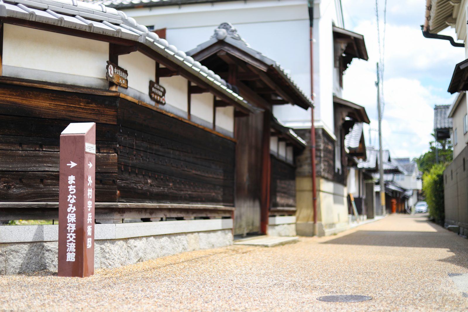 A reddish-brown signpost with an arrow stands beside a dark weathered plank wall and a white-plastered storehouse along a quiet gravel-paved street, with more traditional buildings stretching ahead.