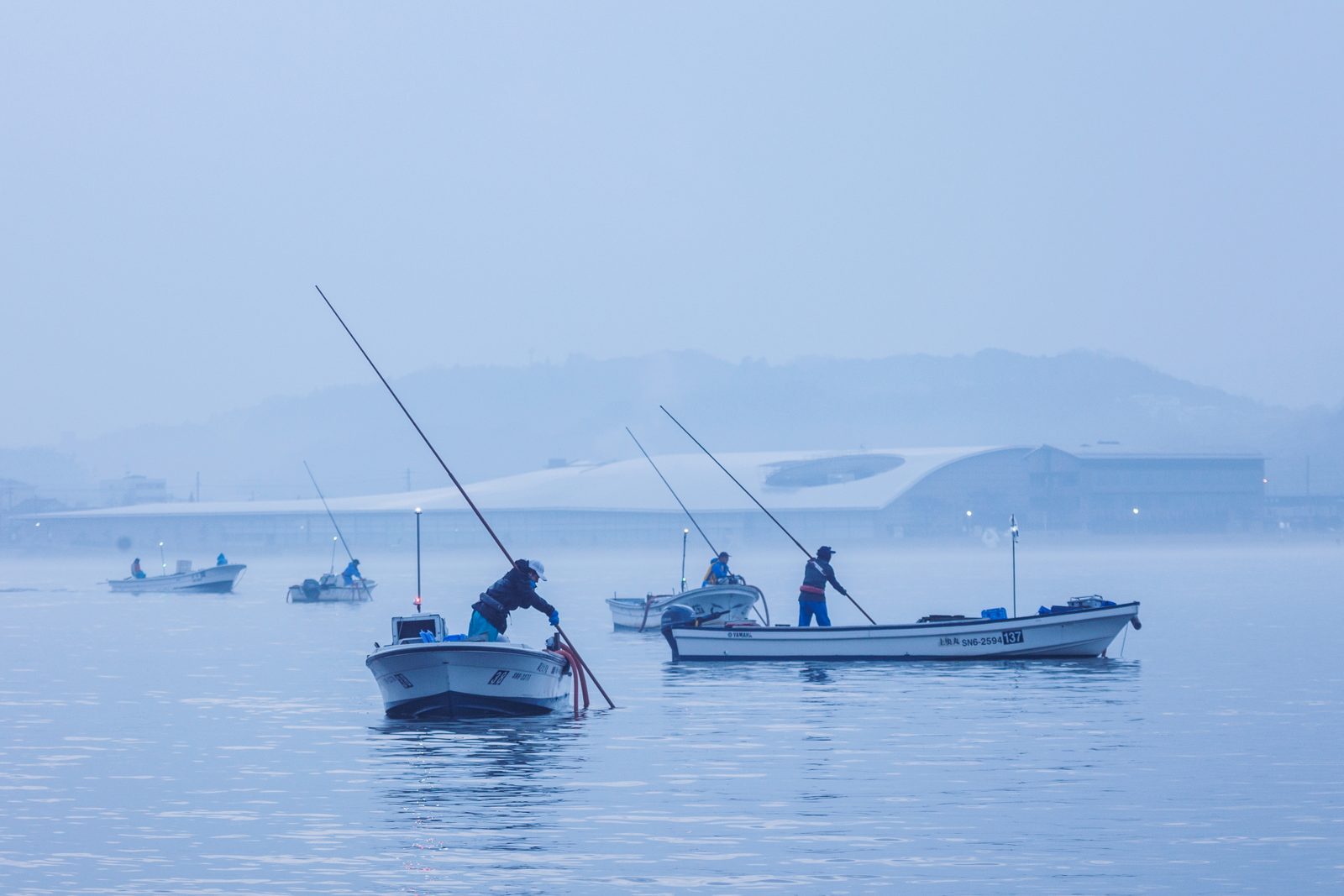 Clam fishers standing in small boats and working long poles on the pale blue, misty water of Lake Shinji