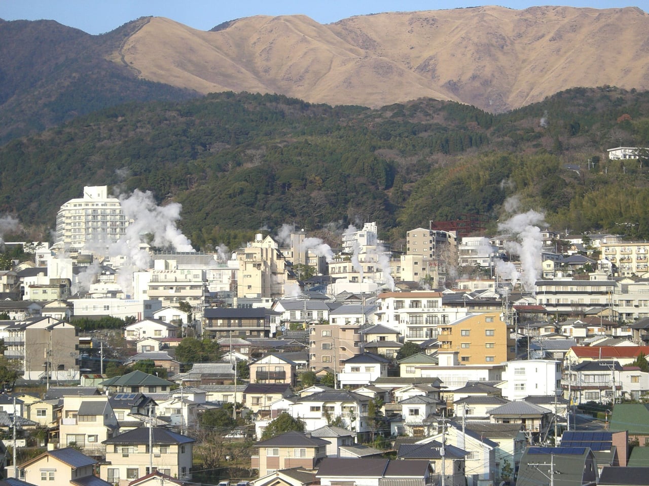 A hillside view of Kannawa Onsen in Beppu, with steam rising between closely packed hot-spring buildings and mountains behind.