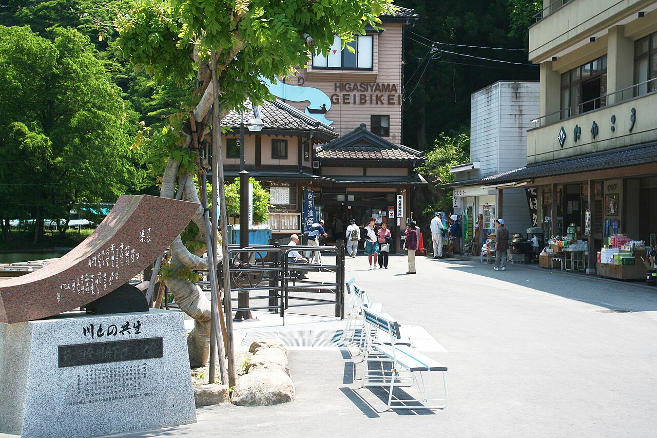 The small square at the Geibikei boat landing, lined with a traditional-style inn, souvenir shops and a stone monument, with visitors strolling in summer light