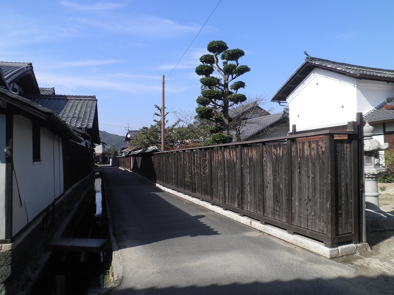 A quiet lane in the Kawanami district of Gokasho, with a dark plank fence on one side, a narrow stone-edged waterway on the other, and a trimmed pine against a blue sky.