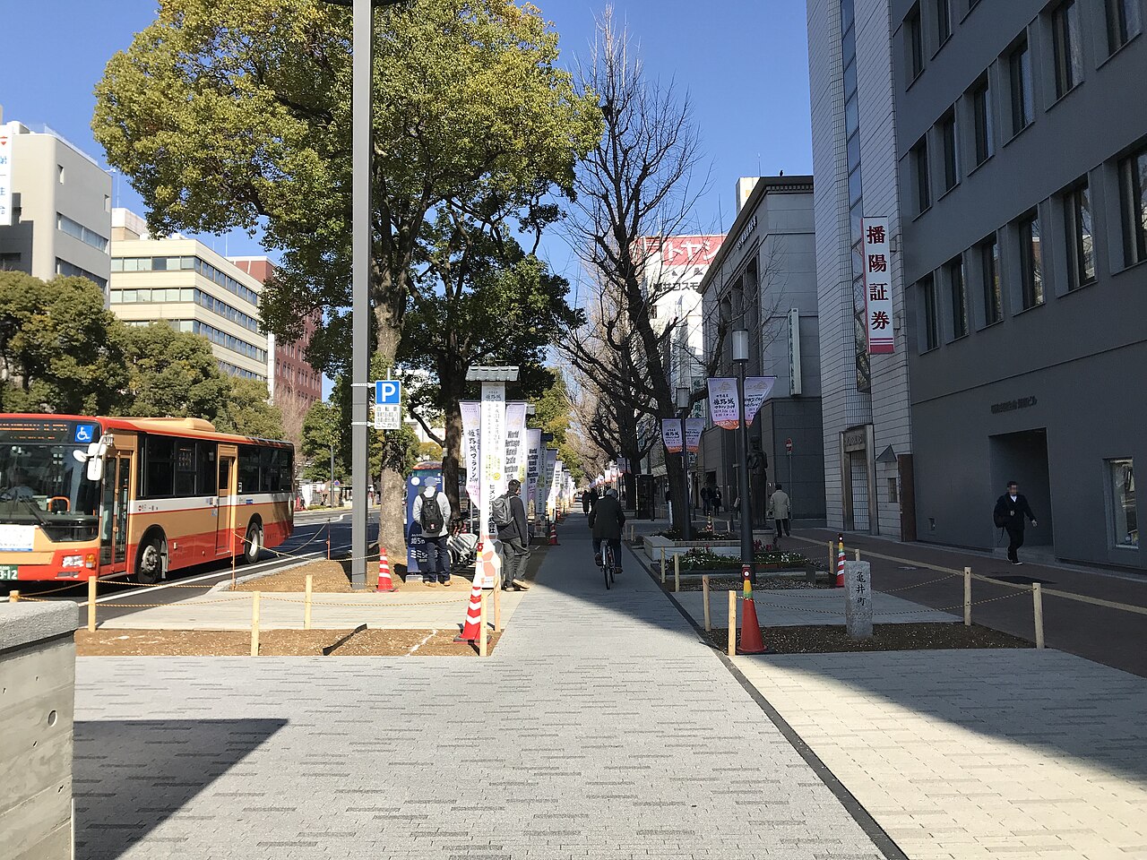 A wide paved sidewalk beside a street lined with trees, banner flags and office buildings, with an orange bus at the left.