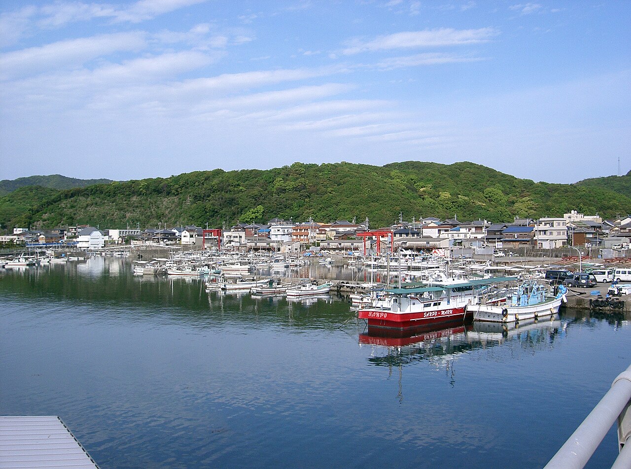Fishing boats moored in Kada harbor with rows of waterfront houses at the foot of green hills.