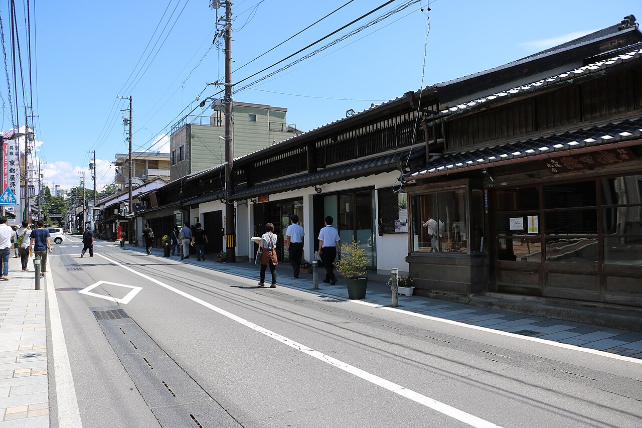 A quiet street of old tile-roofed merchant houses along the former Hokkoku Road in Komoro, with a few people walking on the pavement under a blue sky.