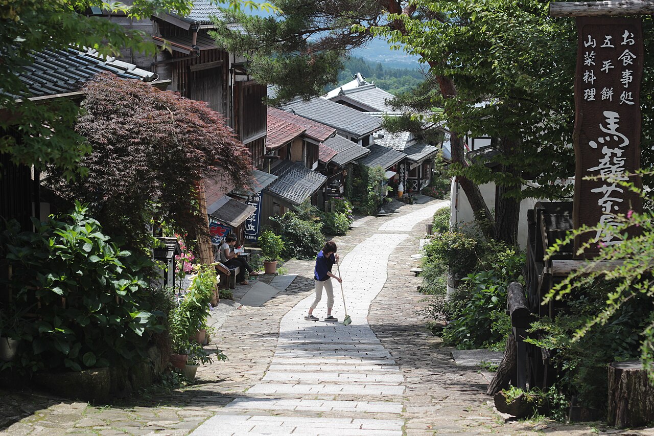 A stone-paved street sloping uphill between old wooden buildings and maple trees in Magome-juku, with a lone walker and a signboard on the right