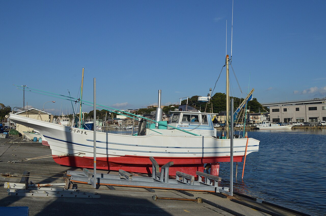 A white-and-red fishing boat resting on a slipway beside blue water in the fishing port of Misaki, with warehouses in the background