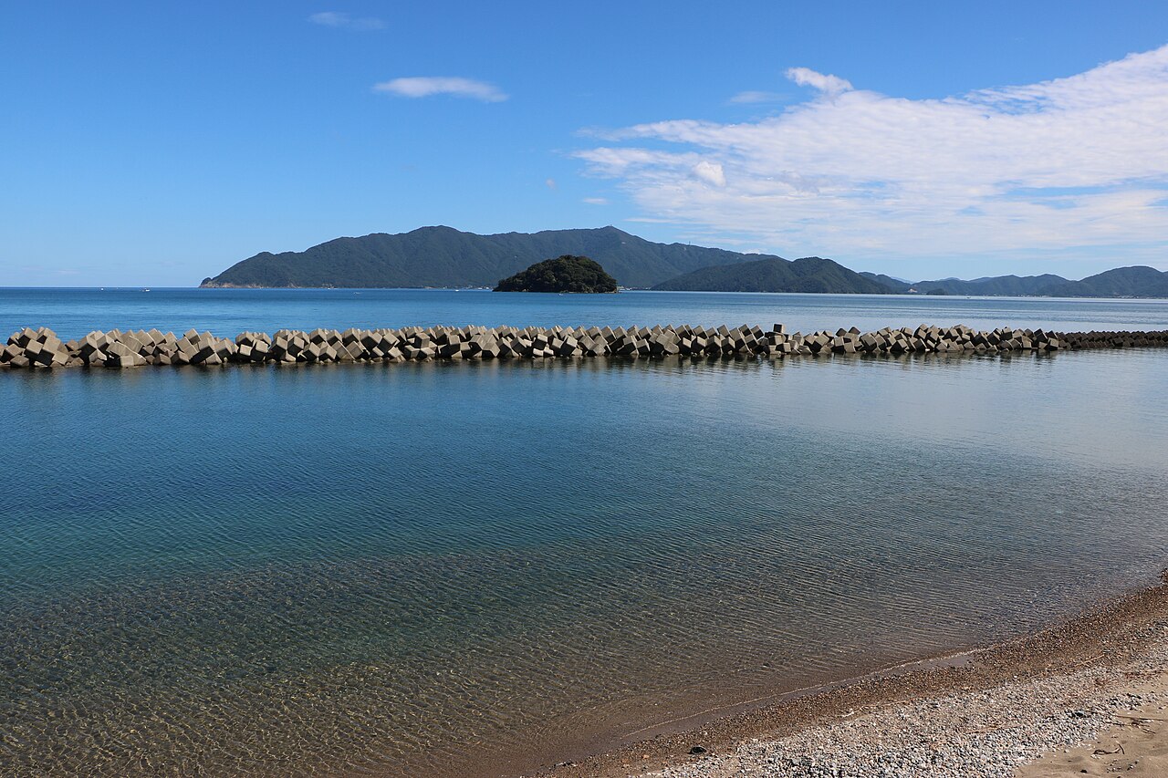 Calm shallow water and a breakwater of concrete blocks on Obama Bay in Fukui, with the small wooded island of Aoshima and hills beyond under a blue sky