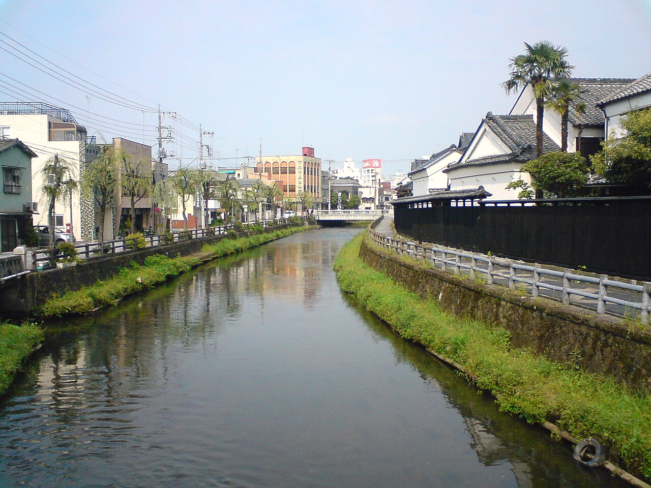 The calm Uzuma River running straight through central Tochigi, with grassy banks, white storehouses and a long black fence on the right and willows along the far bank
