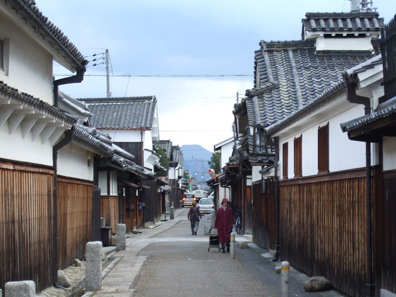 A narrow street between white-walled storehouses and wooden fences in Tondabayashi's Jinaimachi, with two people walking and a hill in the distance