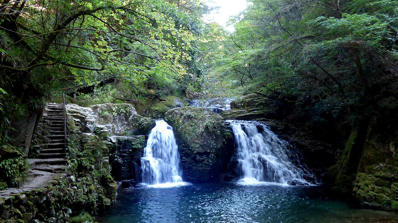 Two small waterfalls dropping into a deep blue-green pool, with a stone staircase and handrail on the left and trees overhead