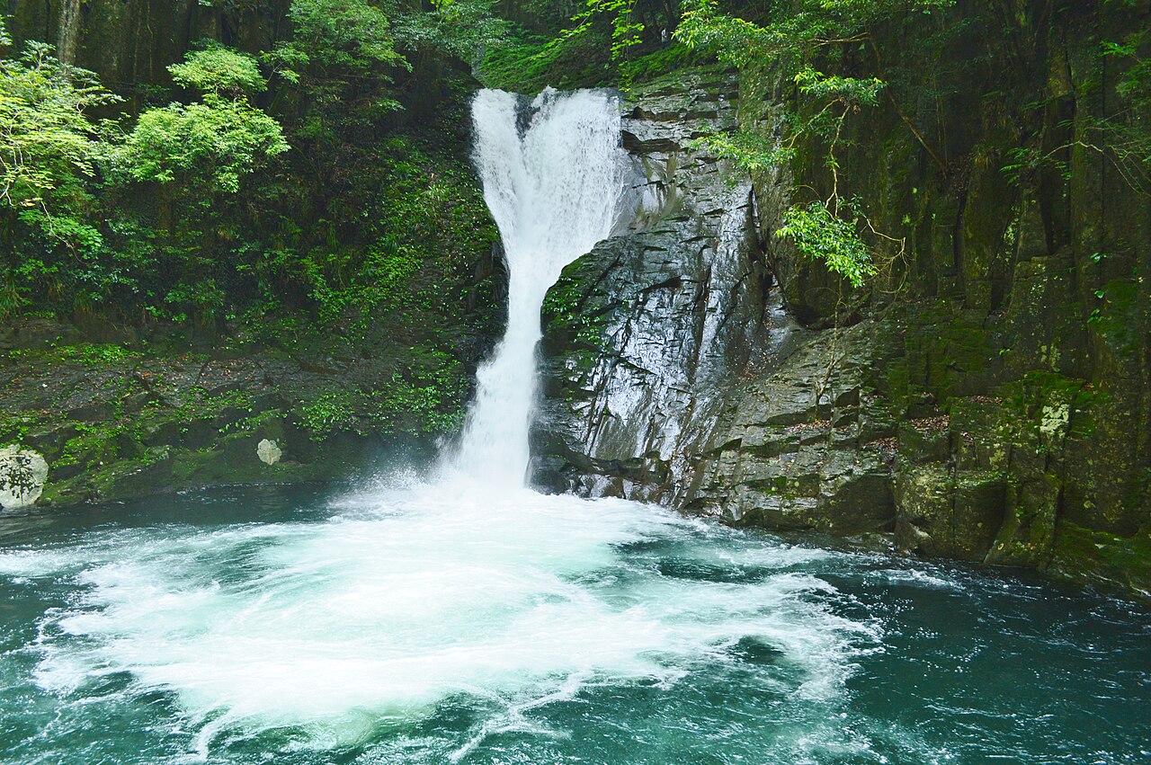 A narrow waterfall dropping beside a dark rock face into a churning, white-foamed pool, with green trees above