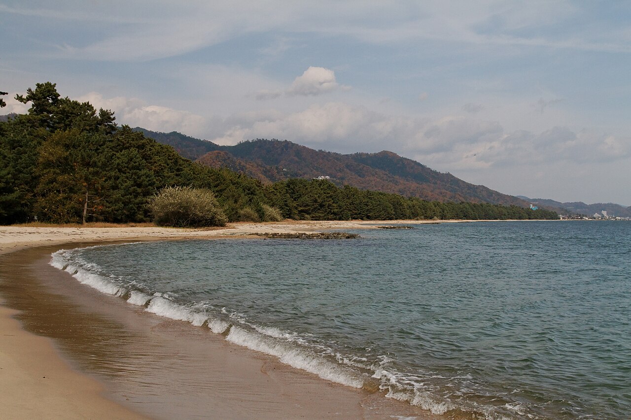Quiet sandy beach with gentle waves and pines along the Amanohashidate sandbar, with hills across the bay