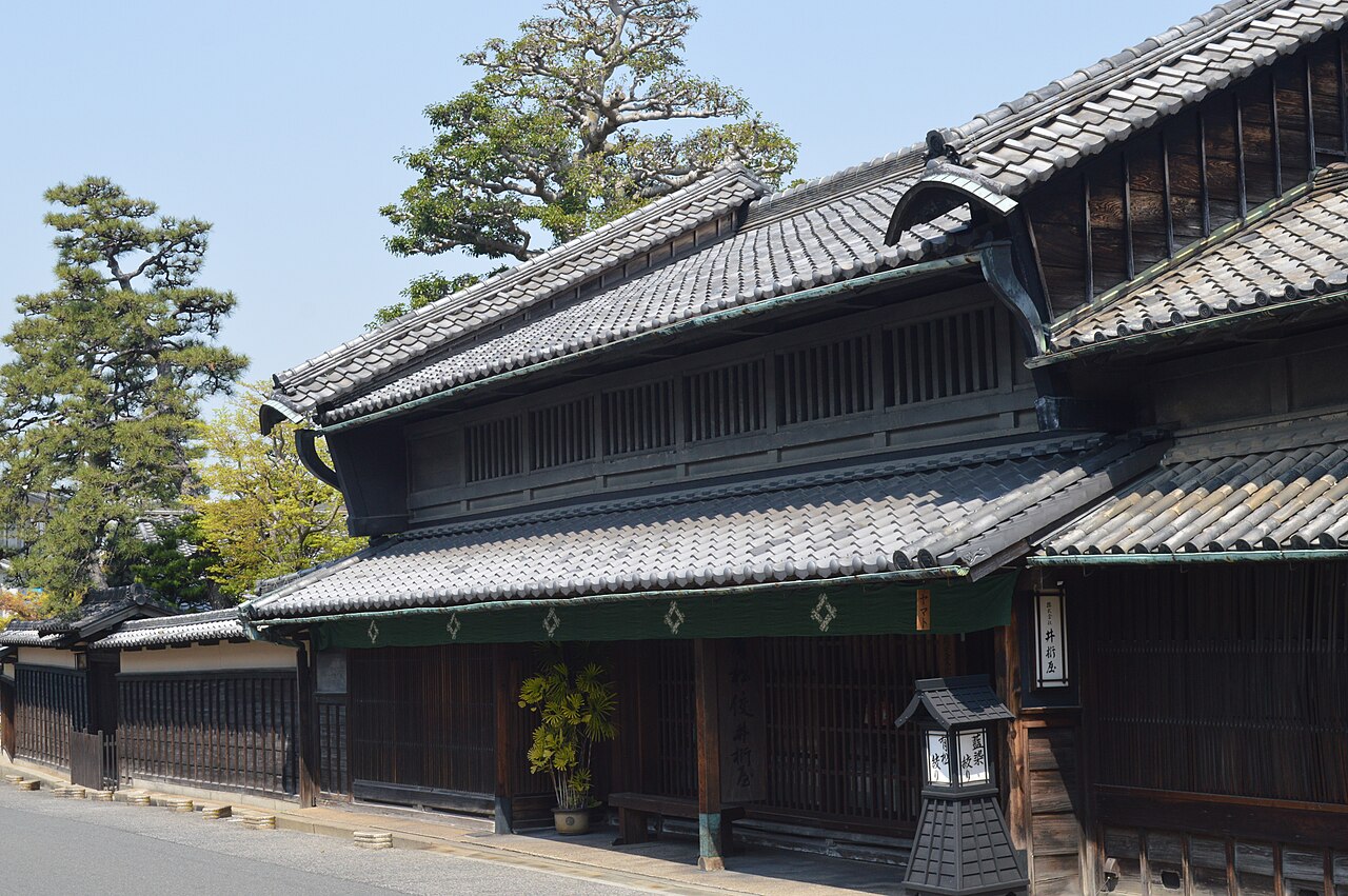 A two-storey old merchant house with layered tiled roofs, dark wooden latticed walls, a green half-curtain and a small lantern at the entrance, beside a narrow street.