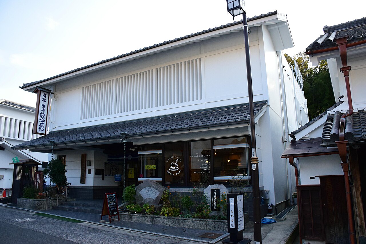 A white-walled two-storey building with a vertical signboard reading Arimatsu Narumi Shibori Kaikan, a tiled eave over the entrance, a street lamp and small shrubs in front.