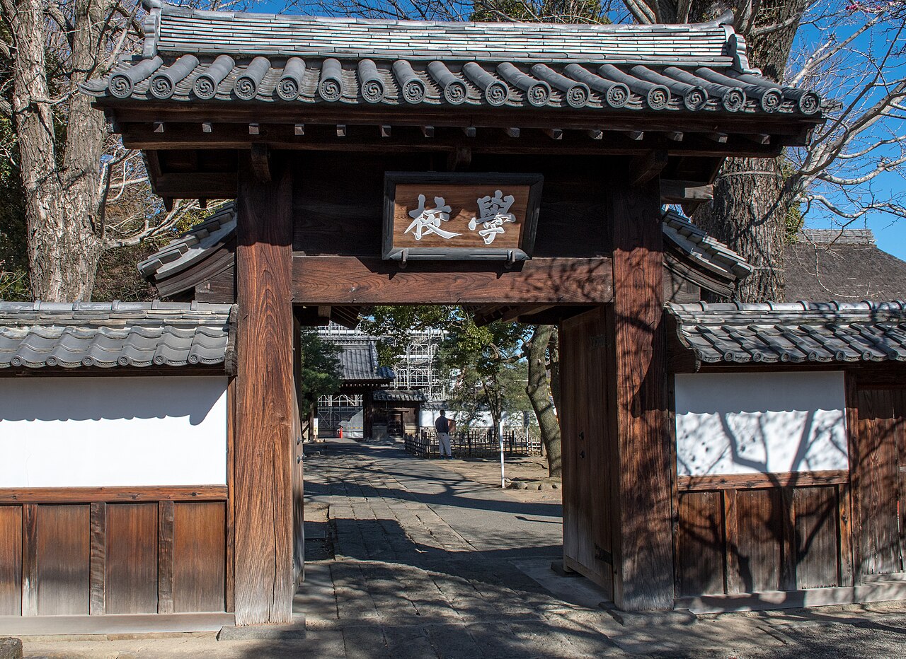 The wooden school gate of Ashikaga Gakko, with its tiled roof, the plaque reading Gakko, and the stone path leading inside