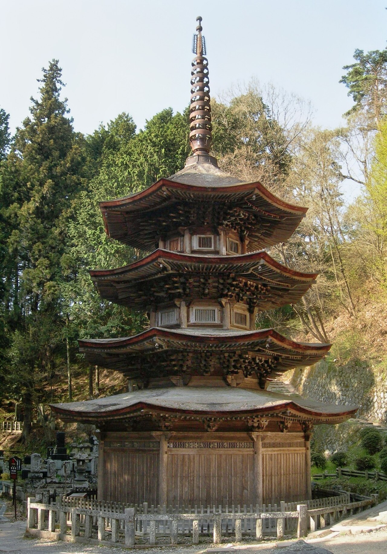 A wooden three-storied octagonal pagoda with layered curved eaves and a tall finial, standing among trees beside a low stone fence at Anraku-ji temple.