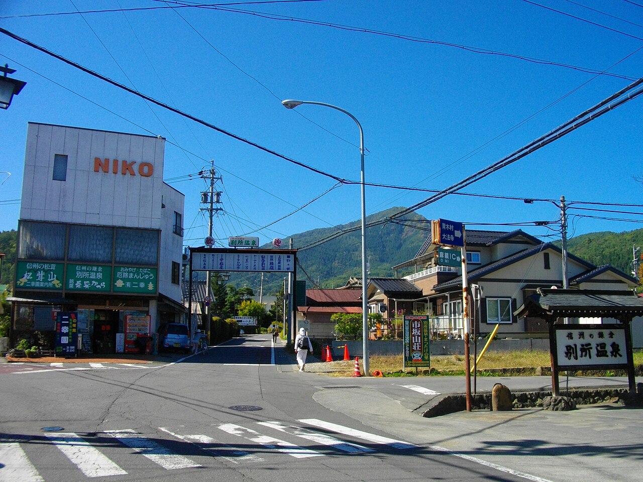 A crossroads at the edge of Bessho Onsen with a small gate sign above the road, a wooden signboard reading Bessho Onsen, and wooded mountains behind the low buildings.