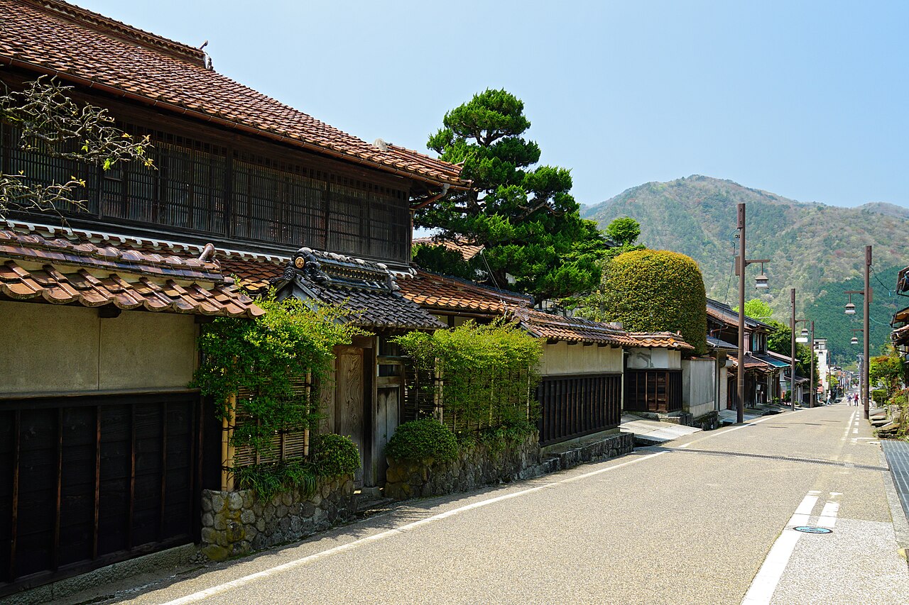 Quiet street in the Chizu post town with tile-roofed wooden merchant houses, a tall pine and wooded hills under a clear sky