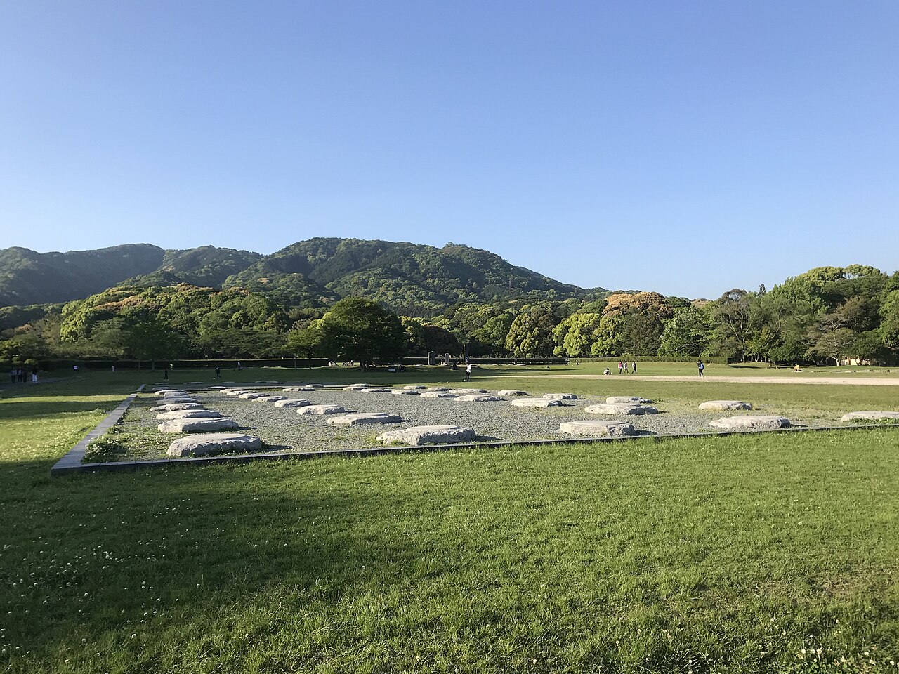 Foundation stones lined up on the wide grassy site of the Dazaifu government office ruins, with a range of mountains rising behind.