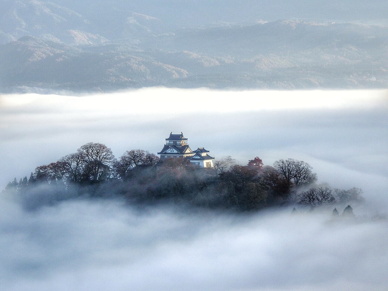 Echizen-Ono Castle with its small white keep on a wooded hilltop rising above a thick sea of clouds, with layered mountain ridges behind.