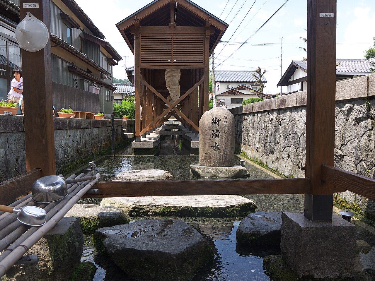 Oshozu spring in Ono, a clear stone-lined pool with stepping stones, a small wooden roofed well house and a stone wall beside old houses.