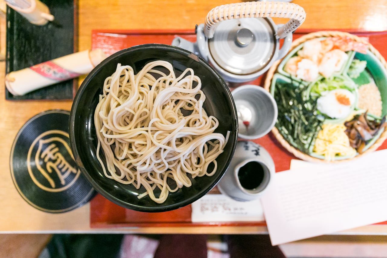 An overhead view of soba noodles in a lacquer bowl beside a dish of toppings including shrimp tempura and omelet strips, with a teapot and small cups on the tray.