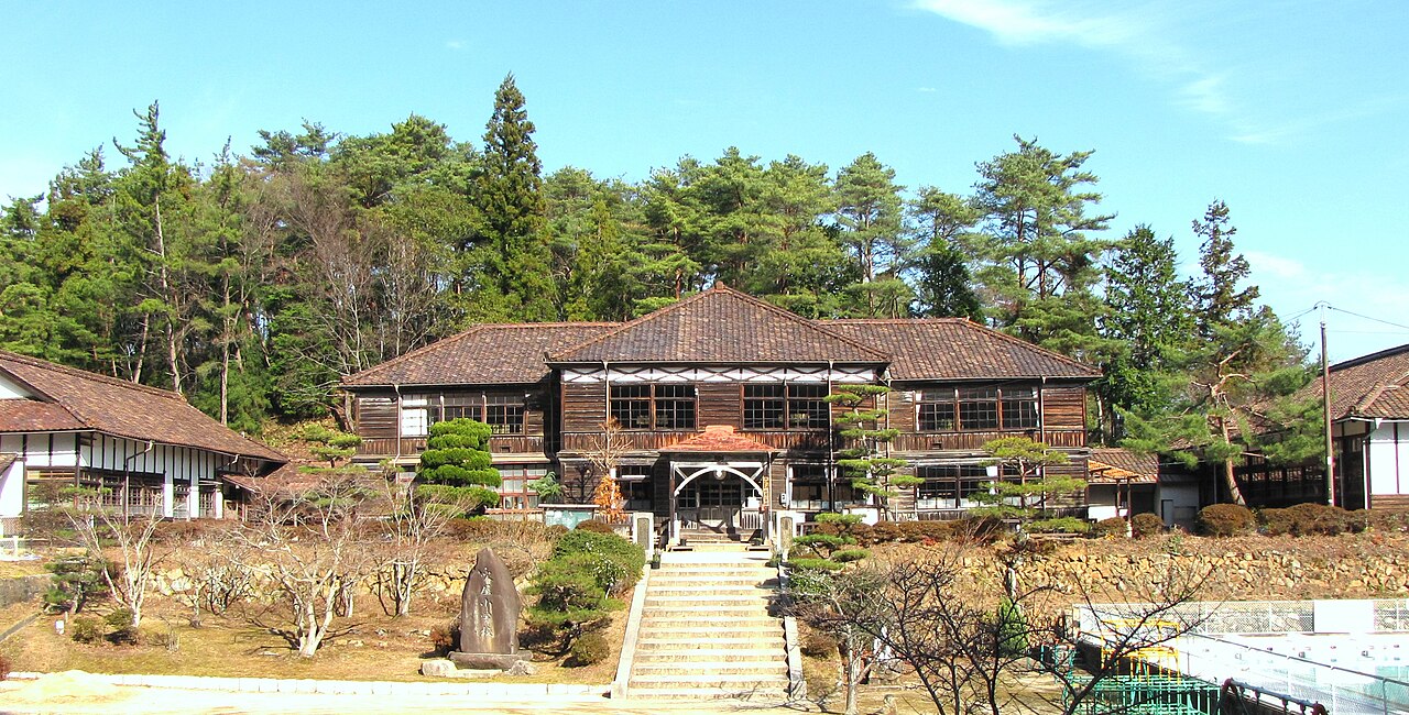 The old wooden Fukiya Elementary School building with a tiled hip roof and entrance stairs, set among pines on a hillside