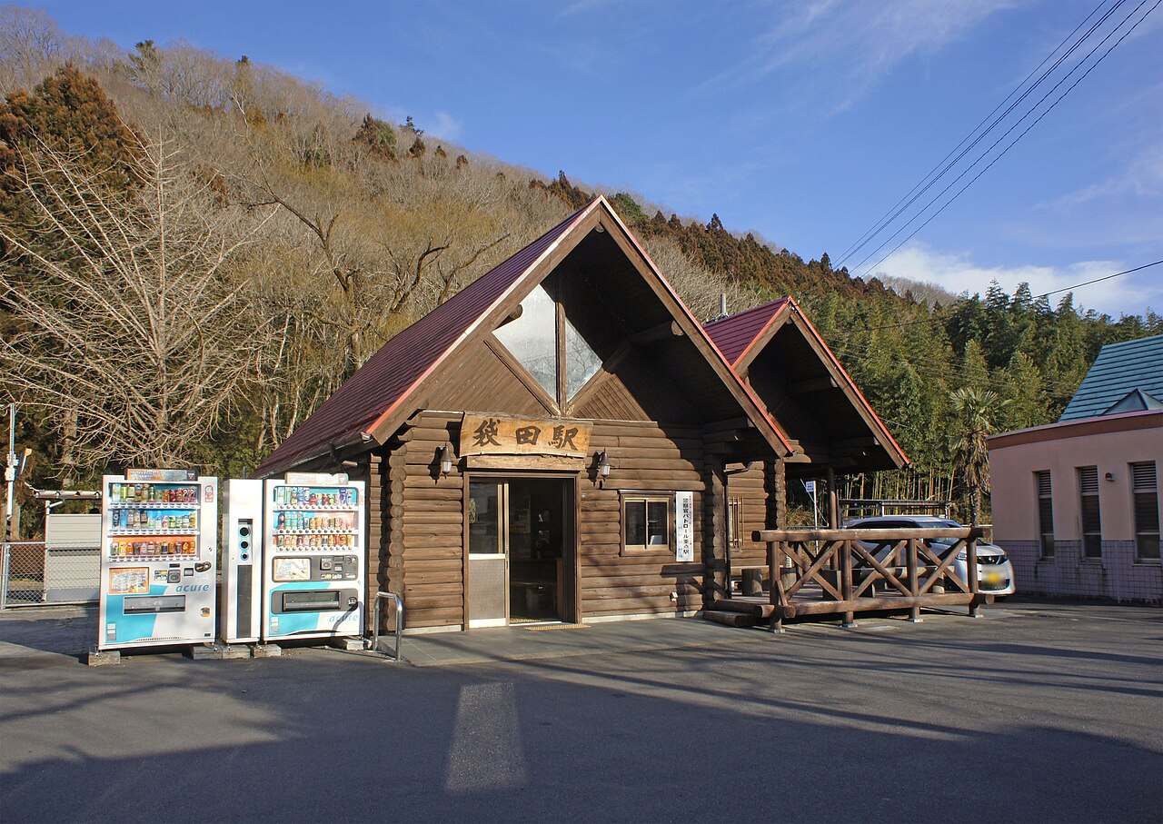 A small log-built station building with a steep gabled roof and a car park in front, with wooded hills behind it.